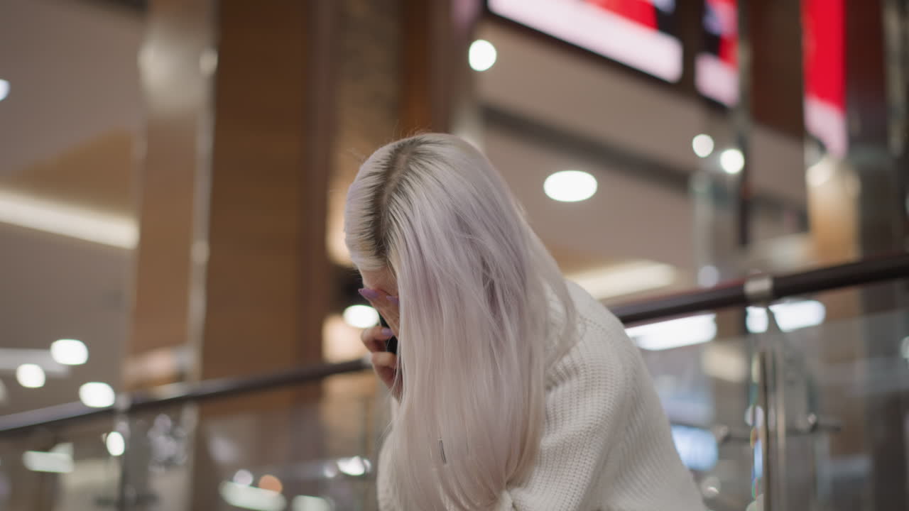 guest seated on mall bench speaking on phone while shaking head and covering mouth thoughtfully, dressed in white sweater, jeans and watch, glass railing, blurred lights and black purse beside her