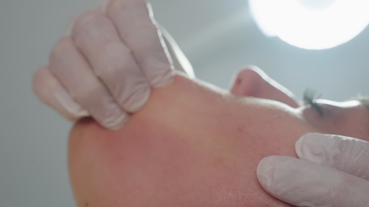 Close up of recipient lying face up under lamp with eyes closed and mouth open gently while skin expert wearing gloves dips fingertip inside mouth and massages cheek in serene clinical setting