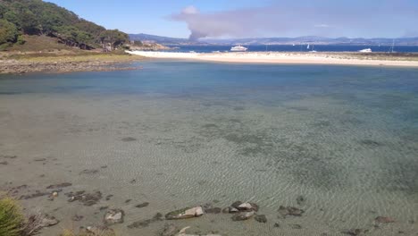 Fire-on-the-horizon-behind-the-coast-with-forest-and-beach-with-luxury-yachts-anchored-on-a-sunny-summer-day,-rolling-crane-shot-upwards,-Cíes-Islands,-Pontevedra,-Galicia,-Spain