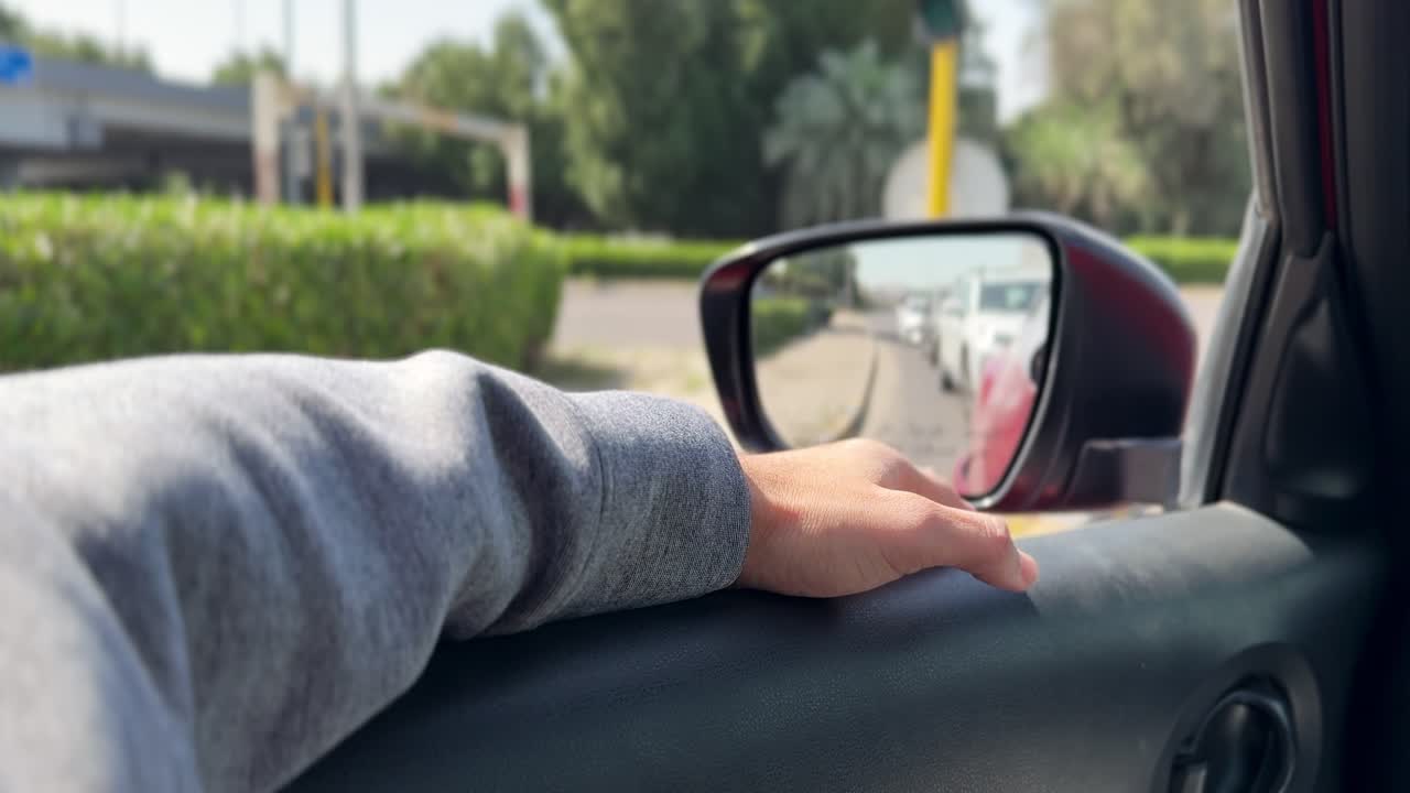 Vista ravvicinata dall'interno della mano di un passeggero appoggiata sulla portiera dell'auto, accanto allo specchietto retrovisore laterale, mentre attende nel traffico urbano durante un pendolarismo diurno luminoso.