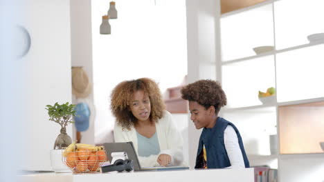 happy african american mother helping her son with homework using tablet in kitchen, slow motion