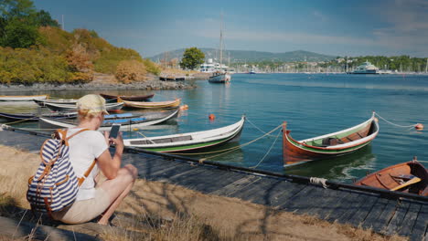 a woman with a backpack sits on the waterfront of the city of oslo in norway near the fishing boats