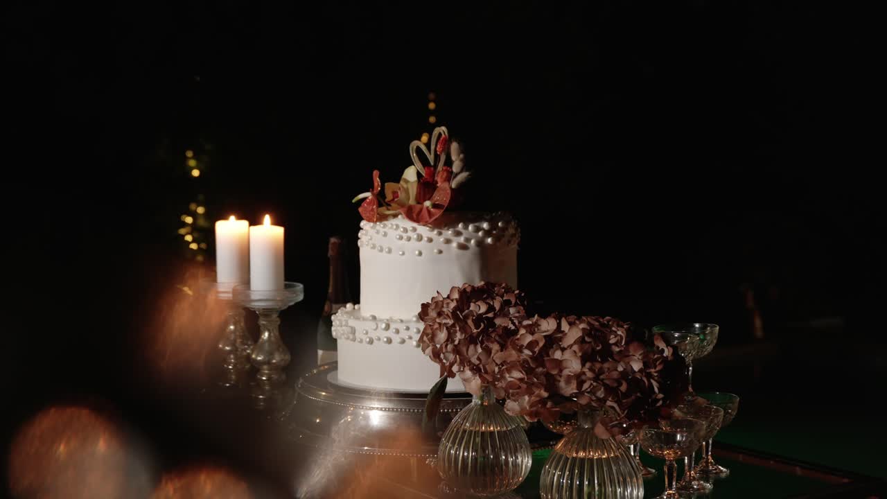 two tiered white wedding cake with sugar pearls and florals lit by candles and surrounded by glass decor