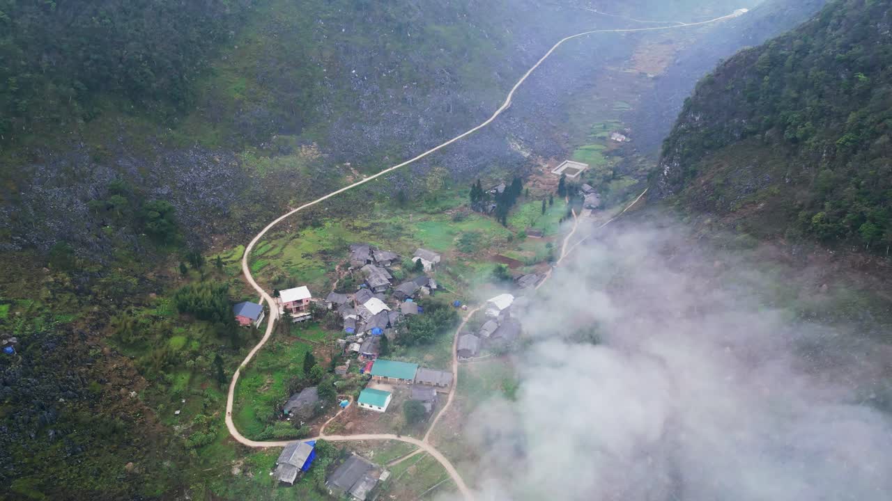 ภาพมุมสูงจากโดรนของหมู่บ้านชนบทบนภูเขาที่ตั้งอยู่ในหุบเขาขรุขระ มีบ้านเรือนกระจัดกระจาย ล้อมรอบด้วยเนินเขาหินและเมฆต่ำที่สร้างบรรยากาศขมุกขมัวแบบที่ราบสูงในเวียดนามตอนเหนือ