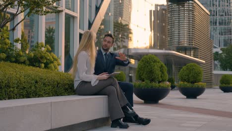 businessman with european blond woman discuss work project ideas sitting outside office building at sunset , man pointing on a side explaining details - full shot, slow motion
