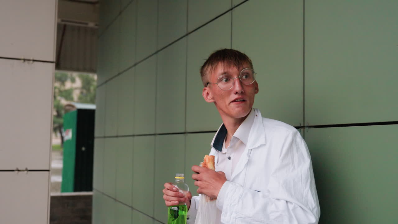 Botany student in white coat standing on porch taking break, holding pastry and sipping juice from bottle, leaning against wall in calm urban outdoor setting with relaxed mood and casual posture