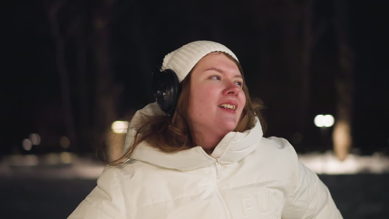 Portrait view of beautiful girl spinning around in snowy park at night, wearing white winter coat and headphones, eyes closed in joyful expression, surrounded by lights in peaceful outdoor setting