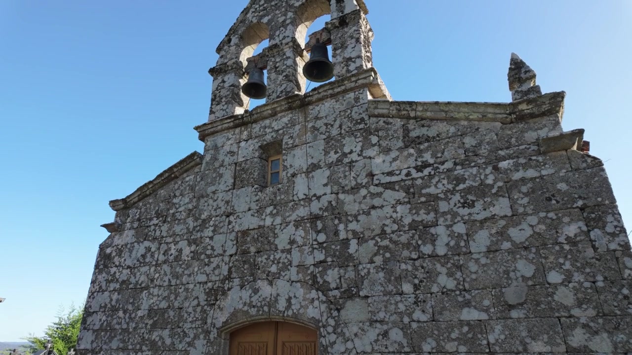 Vista frontale dettagliata della chiesa di San Juan de Paradela de Abeleda a Porqueira, Ourense, che mostra la sua facciata rustica in pietra e il portale in legno nel paesaggio rurale della Galizia, Spagna