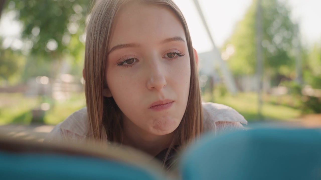 Primo piano di una giovane donna che legge un libro aperto in una luce diurna soffusa con uno sfondo di parco sfocato. La donna indossa un top di pizzo di colore chiaro e cuffie nere intorno al collo, concentrandosi sul testo. Ambiente esterno.