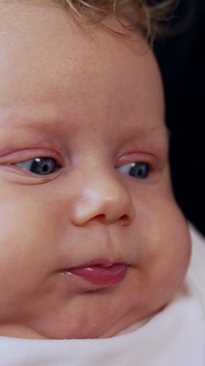 Face of an adorable Caucasian blue-eyed baby with fair hair. Mom's hands hold hands of a child. Close up. Black backdrop. Vertical video