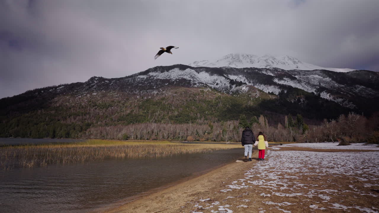 Mother and child walk beside the snowy shoreline of Lago Falkner waving in Neuquén, Argentina, with a bird nearby and snow-capped Andean peaks rising in the background, static camera, real time