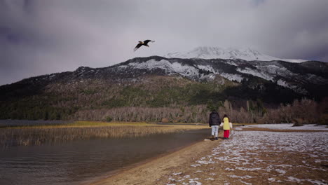 mother and child walk beside the snowy shoreline of lago falkner waving in neuquén, argentina, with a bird nearby and snow-capped andean peaks rising in the background, static camera, real time