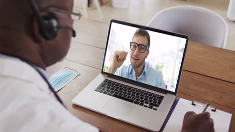 African-american-male-doctor-wearing-phone-headset-taking-notes-while-having-a-video-call-on-laptop