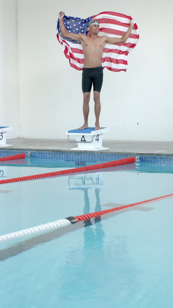 Vertical video: Swimmer holding American flag standing on starting block at indoor pool