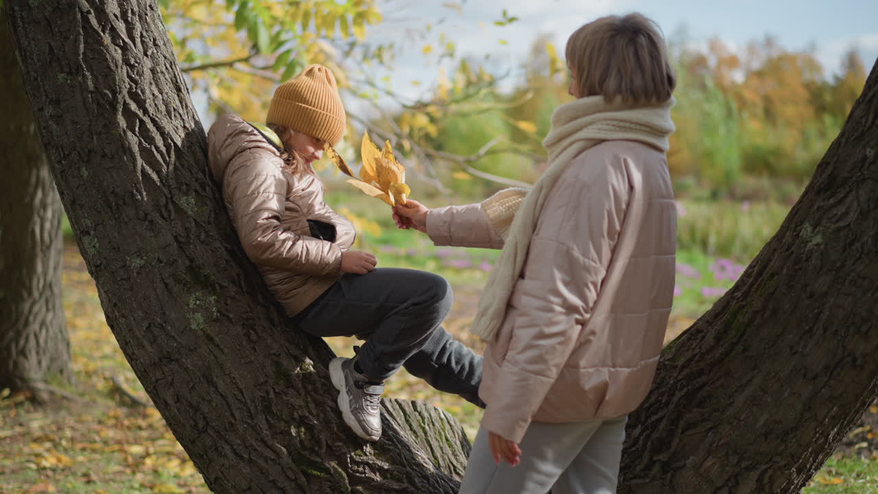 kid rests on tree trunk in autumn park as mother approaches holding yellow leaves and kisses daughter on cheek creating warm tender family moment among fall foliage amid vibrant autumn scenery