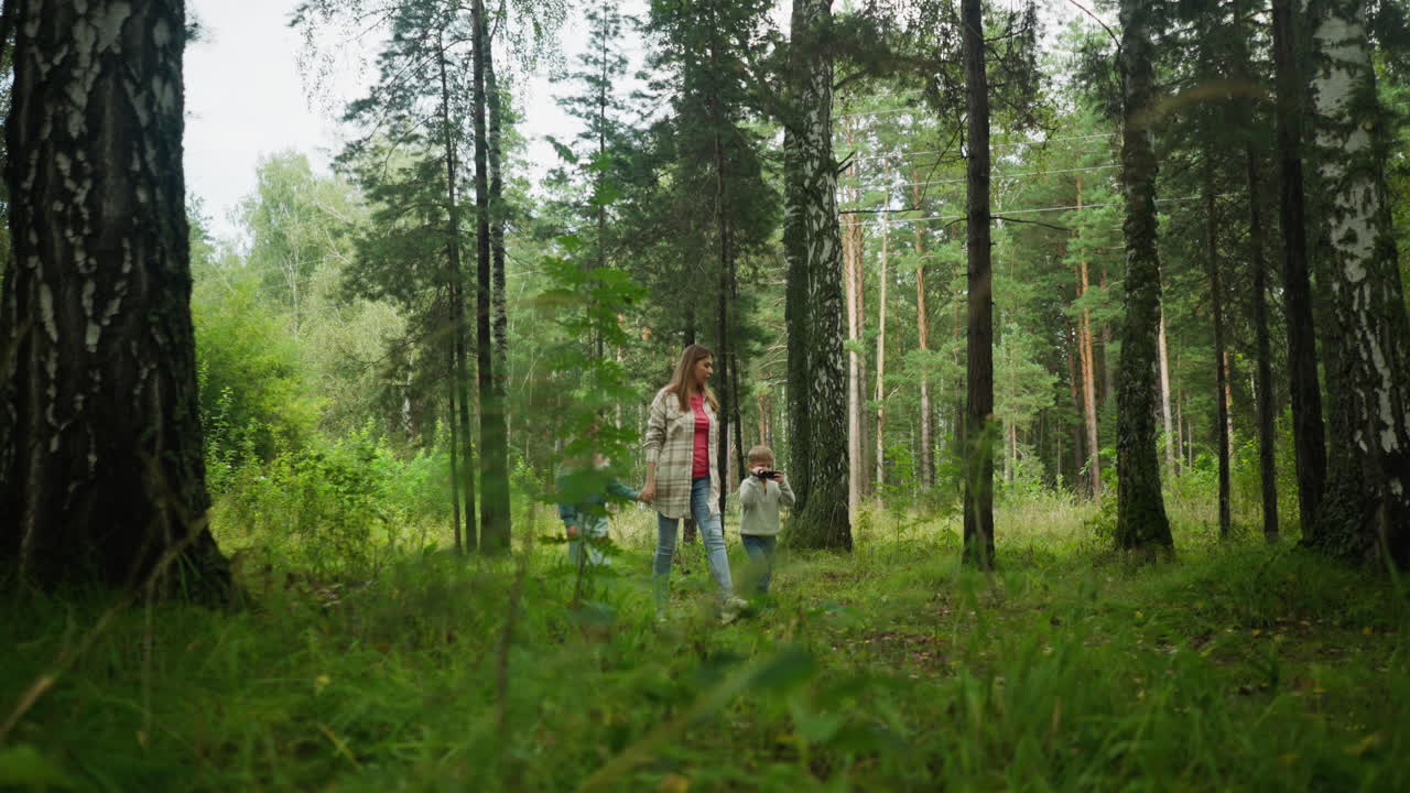 Mother points out something in forest while walking with her kids, as daughter bends down to pick object from ground, surrounded by tall trees, thick greenery