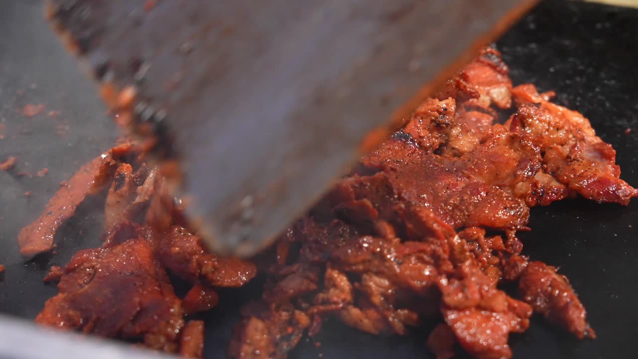 Tampilan jarak dekat POV daging al pastor mendesis yang dimasak dengan spatula dan pisau di atas panggangan jajanan kaki lima Meksiko yang panas, menyoroti aroma, tekstur, dan pengalaman rasa yang gurih serta lezat.