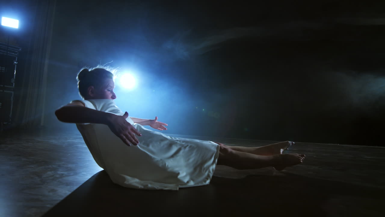 Modern dance woman in a white dress dances a modern ballet jumps makes rotation on the stage with smoke in the blue spotlights