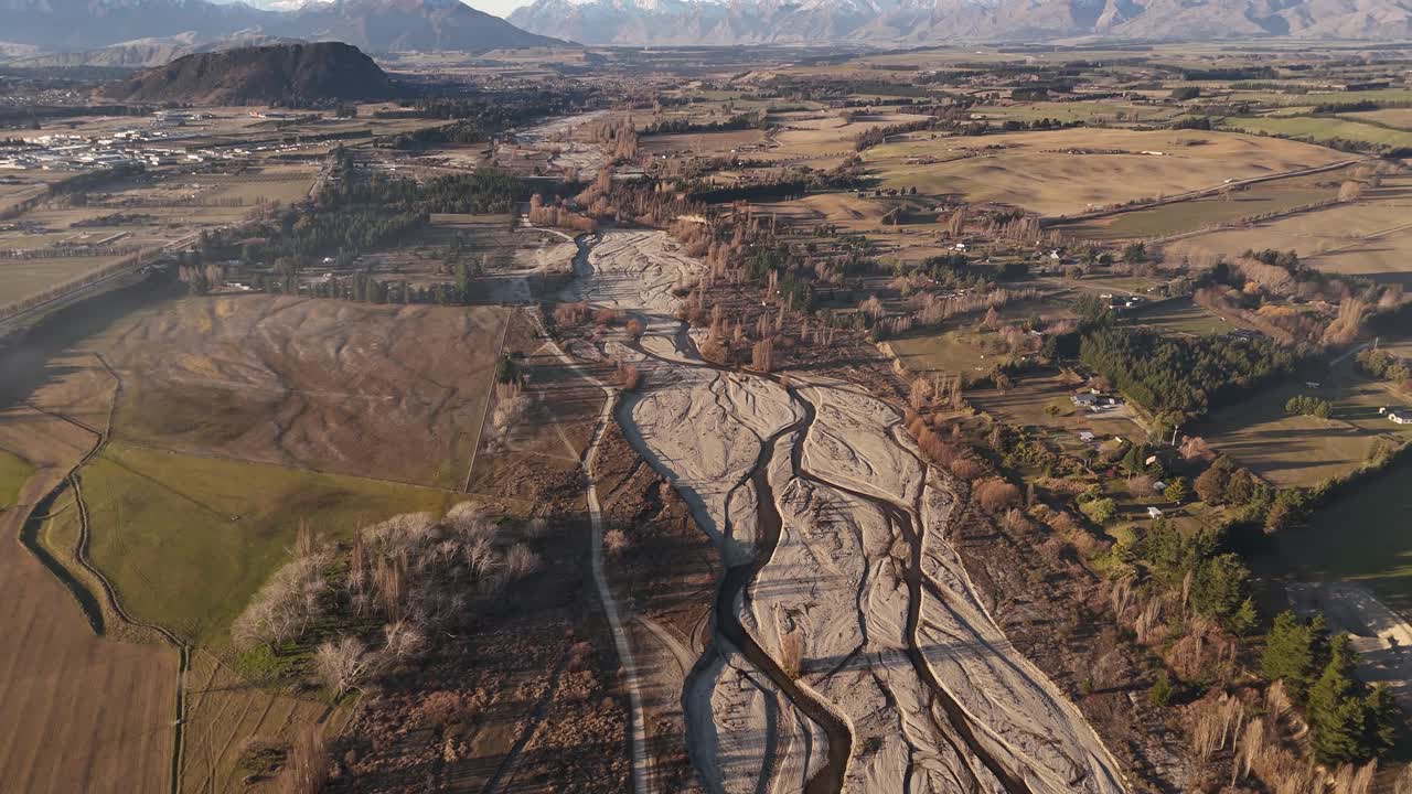 Aerial over the braided, dry riverbed of Wanaka River at sunset, surrounded by brown rugged Southern Alps