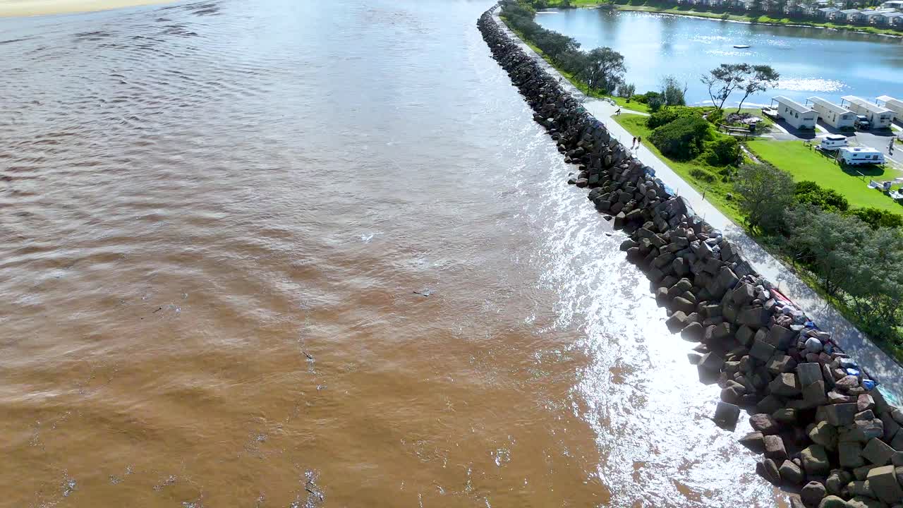 Aerial drone footage over stone breakwater separating brown river and green parkland on sunny day