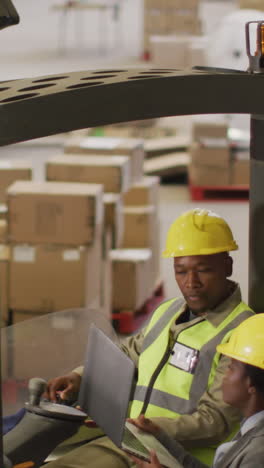 african american male and female workers wearing safety suits and using laptop in warehouse