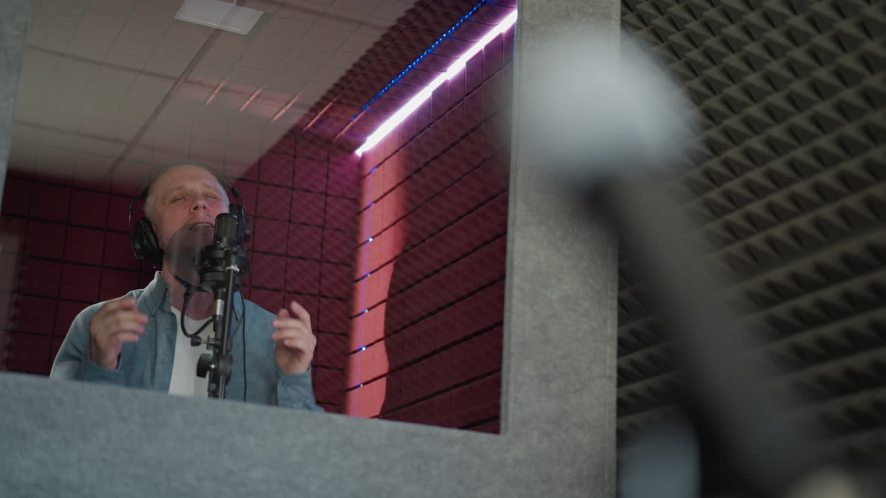 A man passionately sings in a recording studio, wearing black headphones and a blue long sleeve shirt. He performs with his mouth wide open, expressing emotion and intensity