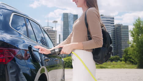 Chica-Cargando-Su-Coche-Eléctrico-En-El-Aparcamiento-Cerca-De-Los-Rascacielos,-Portátil