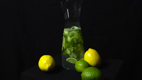 Glass carafe filled with citrus fruit and cucumber water in front of a black studio background. Vibrant yellow and green lime, lemon fruits in front of the glass vessel while footage spins around them