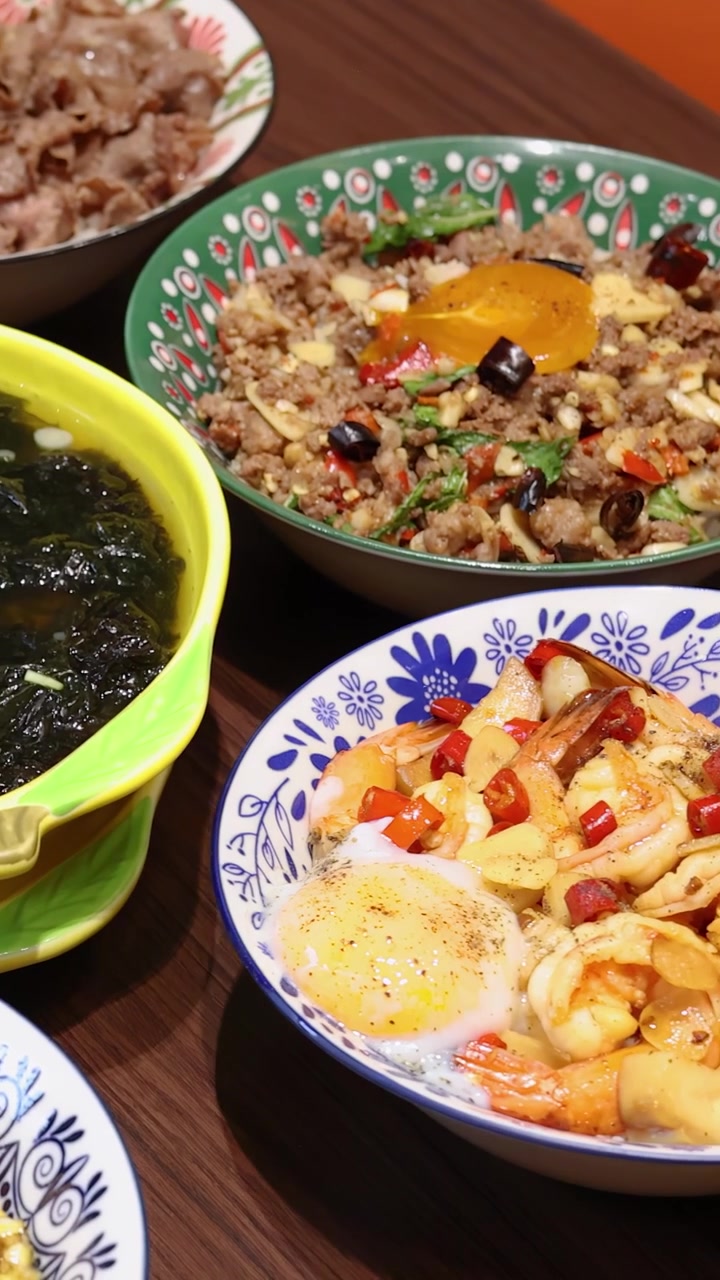 明るい室内照明の下、色とりどりのタイ米料理とスープが木製のテーブルに並べられ、手が一杯の器をよそっている。