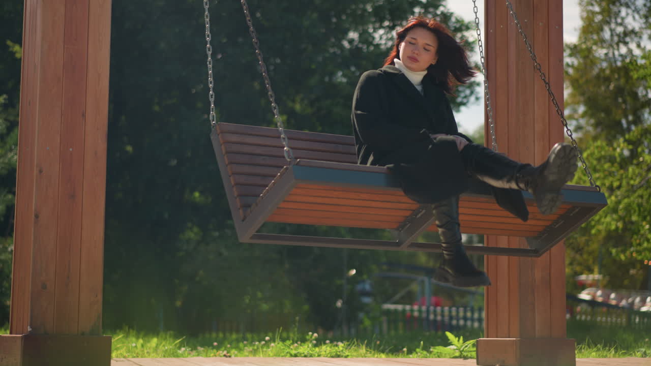 dama con elegante abrigo negro y botas se sienta en un columpio de madera en el parque iluminado por el sol, mirando serenamente a la distancia, teléfono en la silla con fondo de vegetación