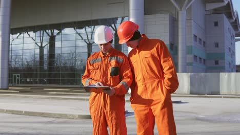 Two-Construction-Workers-In-Orange-Uniform-And-Helmets-Looking-Over-Plans-Together