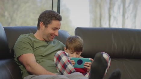 Father-with-son-wearing-pyjamas-sitting-on-sofa-at-home-playing-computer-game-on-handheld-device---shot-in-slow-motion