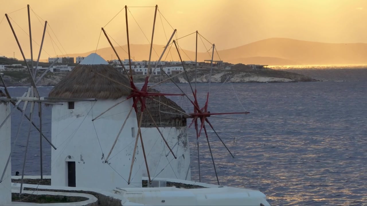 Ripresa statica con drone ad alta angolazione e teleobiettivo che cattura i mulini a vento di Mykonos al tramonto, con un orizzonte dorato e luminoso e un'atmosfera costiera calma.