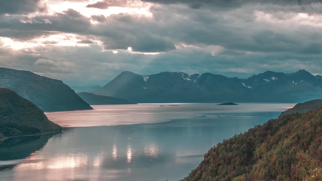Stråler fra den nedgående sol skinner gennem stormskyerne over den fredfyldte fjord i en timelapse-video
