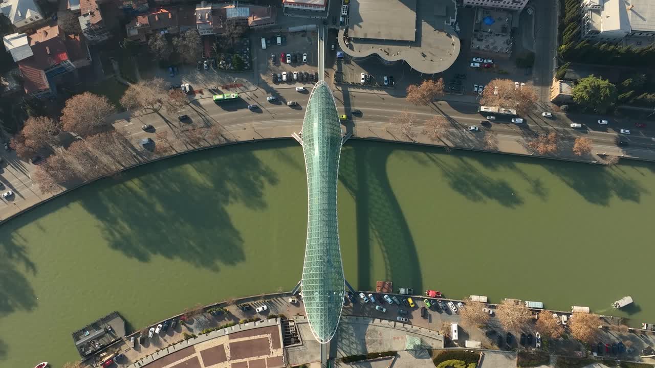 Vertical aerial view showing the Bridge of Peace crossing the Kura River in Tbilisi. The photo emphasizes the bridge’s elegant curves, green water, and surrounding riverbanks on a bright day