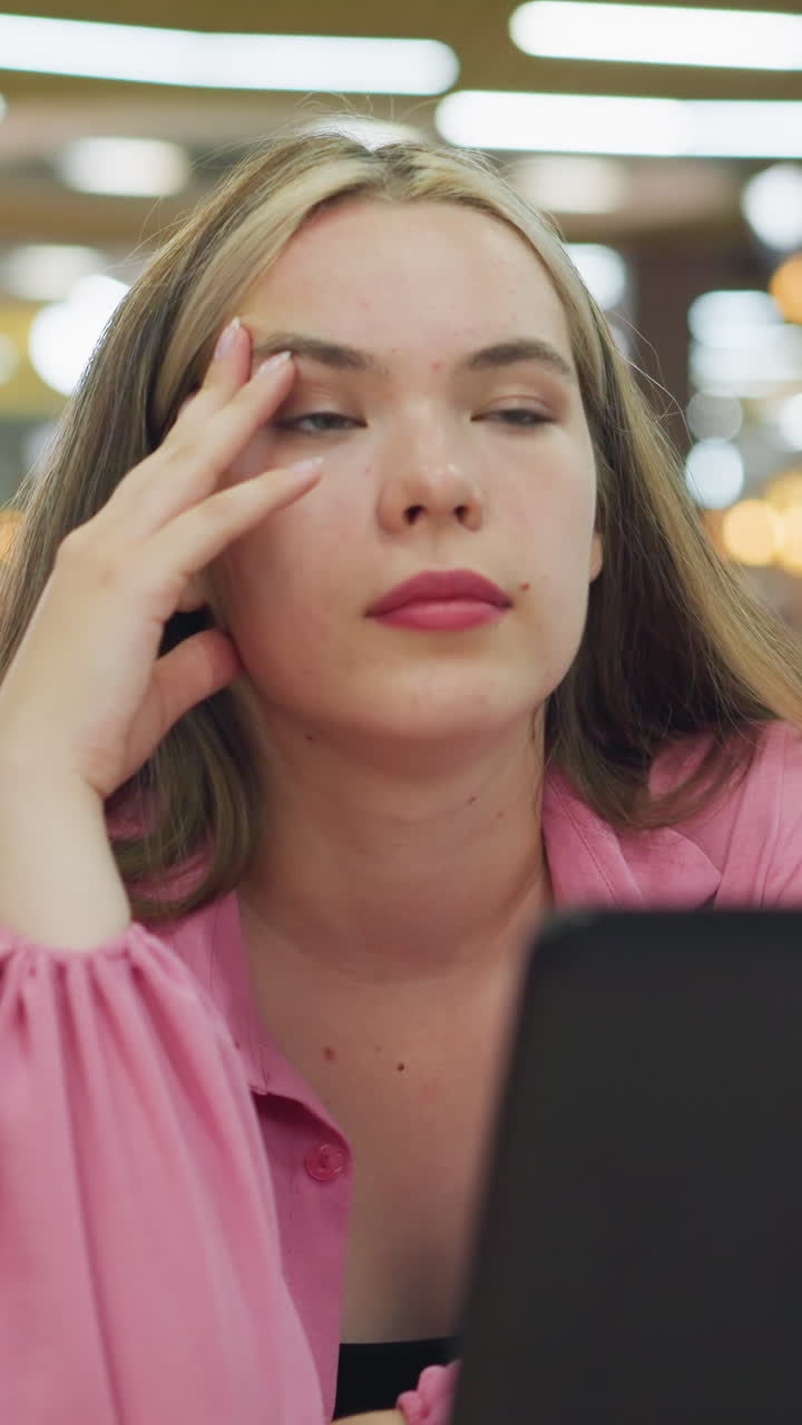 una mujer joven con un vestido rosa se sienta frente a su portátil, colocando su mano sobre su cabeza mientras parpadea y aparece profundamente en el pensamiento, el fondo borroso de un restaurante ocupado y bien iluminado con luz borrosa
