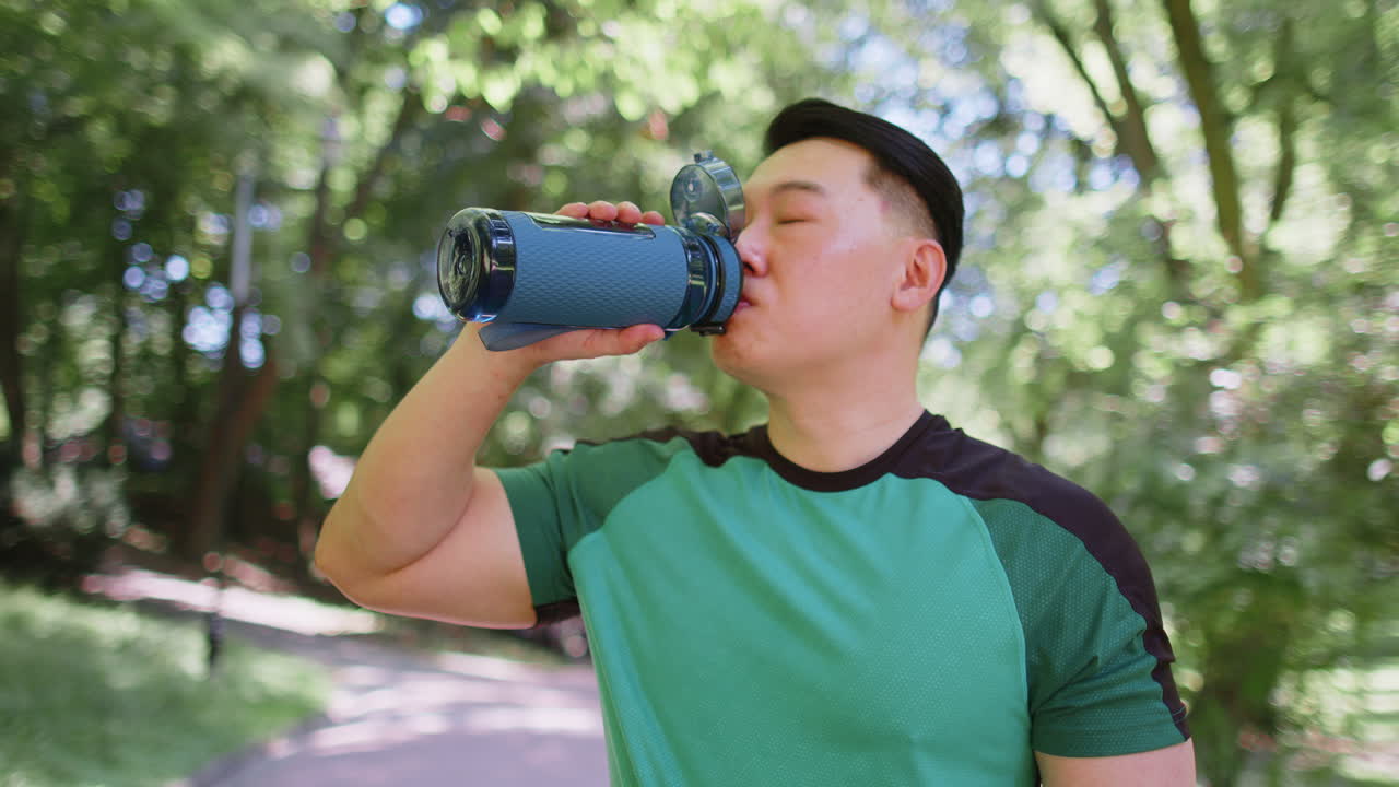 Sporty athletic thirsty chinese asian sportsman runner jogger drinking water after workout in park