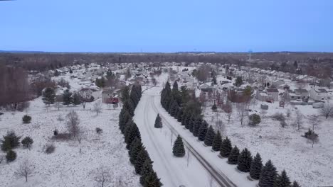 Snowy-small-town-road,-subdivision-entrance-lined-with-pine-trees-,-rural-snow-covered-street-with-a-car-driving-on-a-cloudy-day-with-blue-skies