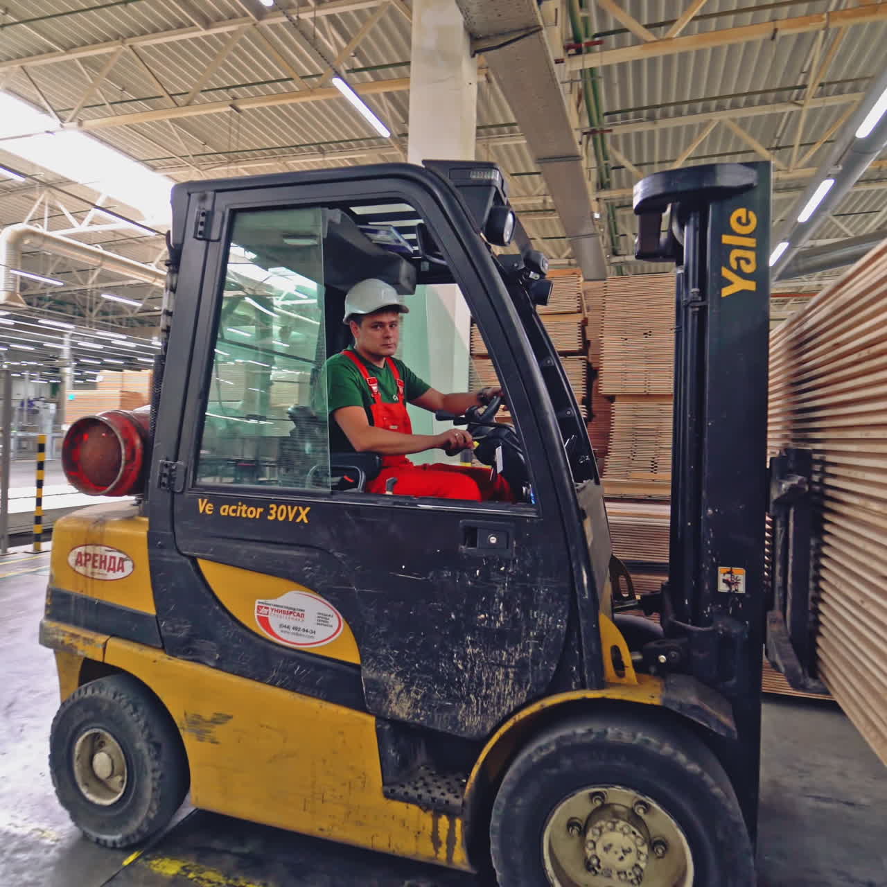 An operator in a white helmet works on an electric loader and carries it a pile of wooden parquet boards to another place in the warehouse. New plant for the manufacture of parquet.