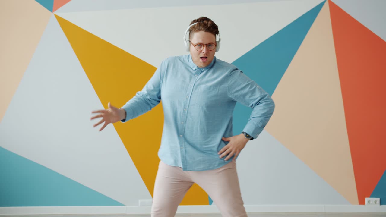 Funny young man in wireless headphones dancing indoors on colorful background