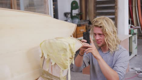 Fabricante-De-Tablas-De-Surf-Masculino-Caucásico-Tomando-Una-Foto-De-Una-Tabla-De-Surf-De-Madera-Con-Su-Teléfono-Inteligente