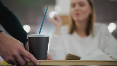primer plano de una mujer borrosa en el fondo leyendo un libro abierto, desayuno servido en bandeja en primer plano, entorno de restaurante moderno con iluminación suave y bokeh vibrante