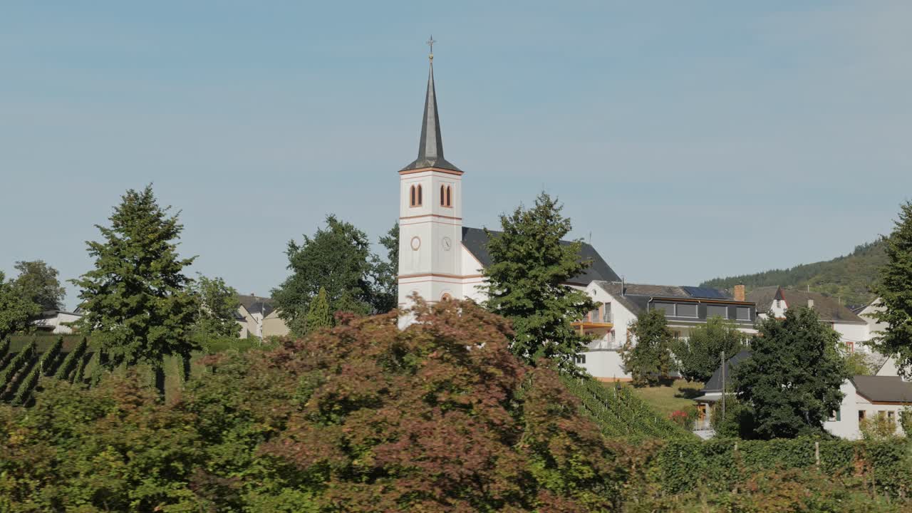 Side view of a church in a small village with vineyards at the river Mosel viewed from a boat with trees passing by. Parallax medium shot.
