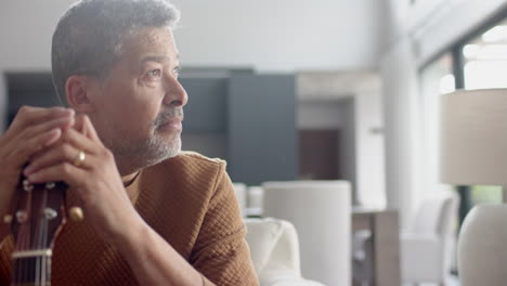 Thoughtful-senior-biracial-man-sitting-holding-guitar-in-living-room,-copy-space,-slow-motion