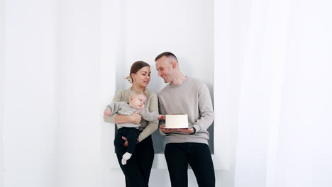 caucasian family in white room. mother holds a little baby and man is holing a cake which the kid tries to get.