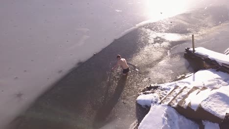 man standing in frozen mountain like wearing shorts no shirt breaks ice with hand to clear hole for ice swimming and polar plunge