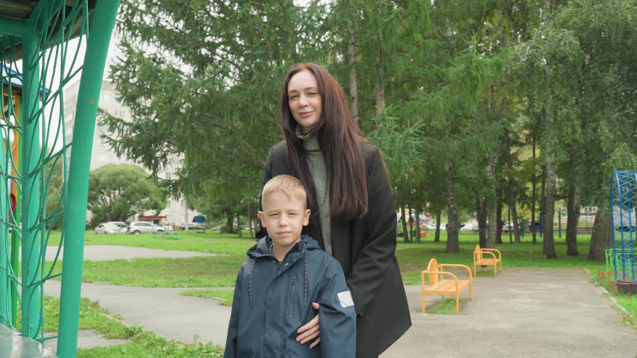 Madre e ragazzo bianco si preparano nel parco della scuola, un dolce addio in un parco giochi con la giacca sistemata, un abbraccio calmante, una silenziosa rassicurazione prima della passeggiata, mattina nuvolosa, verde fogliame e panchina
