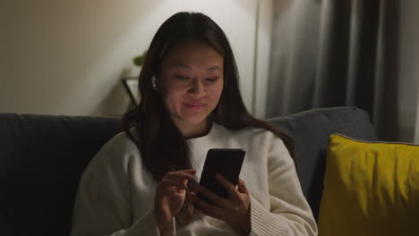 woman spending evening at home on sofa wearing wireless earbuds and streaming music or podcast from mobile phone