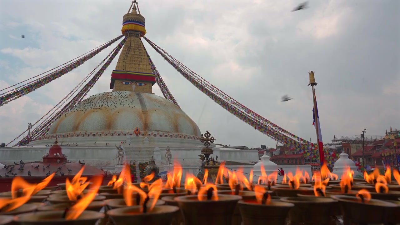Un'inquadratura ampia e statica che mostra lampade a burro tradizionali tremolanti in primo piano con la monumentale Stupa di Boudhanath sotto un drammatico cielo nuvoloso al tramonto a Kathmandu, Nepal.
