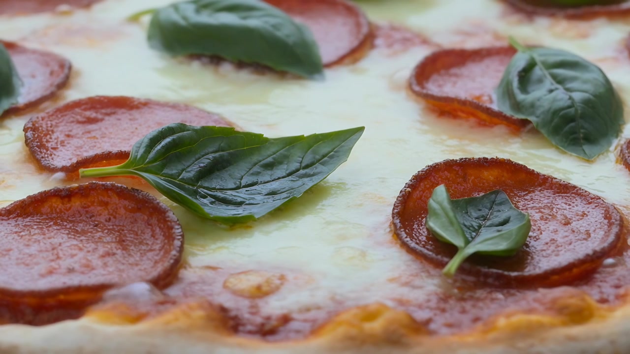 Telecamera che zooma inquadrando una pizza calda al salame piccante con foglie di basilico in studio, mostrando la lucentezza del formaggio.